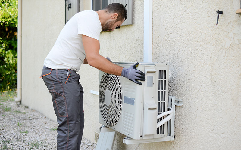A man fixing an air conditioner on the side of a house.